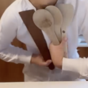 A person in a white shirt is holding several wooden kitchen utensils, including spoons and a flat spatula, against their chest in a kitchen setting.